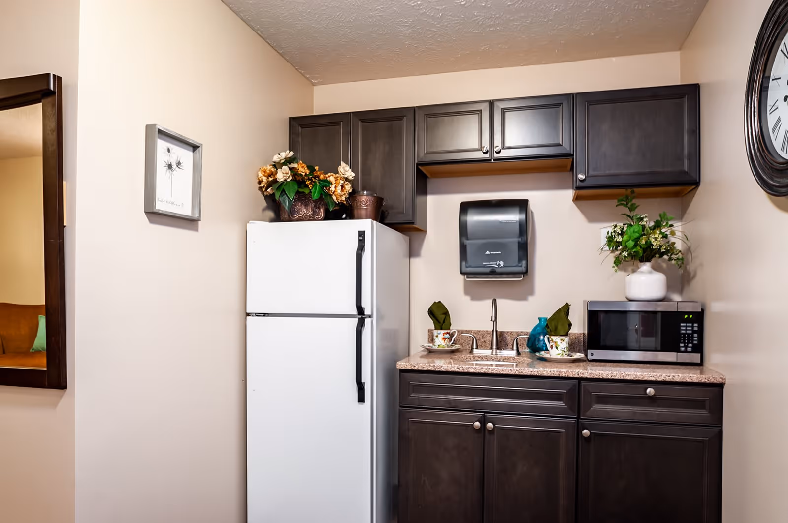 A small kitchen area with dark wood cabinets, a white refrigerator, a countertop with a sink, a microwave, and decorative plants. There are two cups with saucers and green napkins on the counter. A paper towel dispenser is mounted on the wall above the sink, and a large clock is partially visible on the right wall.