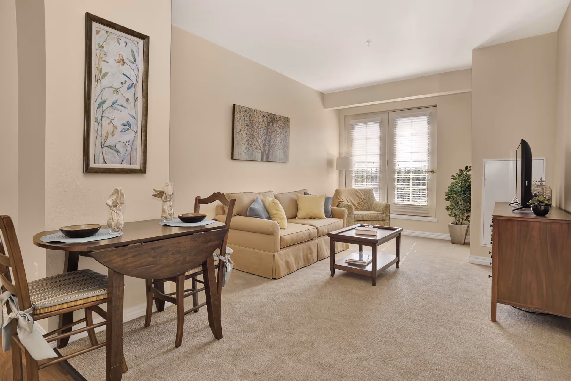 A cozy living room with beige walls and carpet. There is a small wooden dining table with two chairs on the left side, set with bowls and decorative items. In the center, a beige sofa with multiple cushions faces a wooden coffee table. A patterned armchair and a floor lamp are near a window with white blinds. On the right side, a wooden TV stand holds a flat-screen television and a small plant. Two framed artworks hang on the walls, and a potted plant is placed near the window.