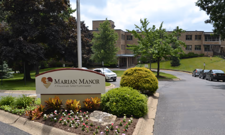 Entrance view of Marian Manor, a Vincentian Senior Community, showing a landscaped area with flowers and bushes in front of a sign, a driveway with parked cars, and a multi-story brick building in the background surrounded by trees and greenery.