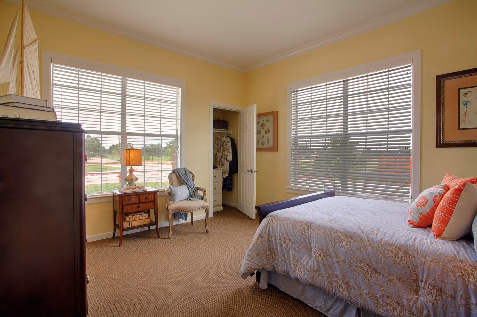 A bright bedroom with two large windows covered by blinds, a bed with patterned bedding and multiple pillows, a wooden nightstand with a lamp, a cushioned chair with a throw blanket, a dark wooden dresser, and an open closet door showing hanging clothes.