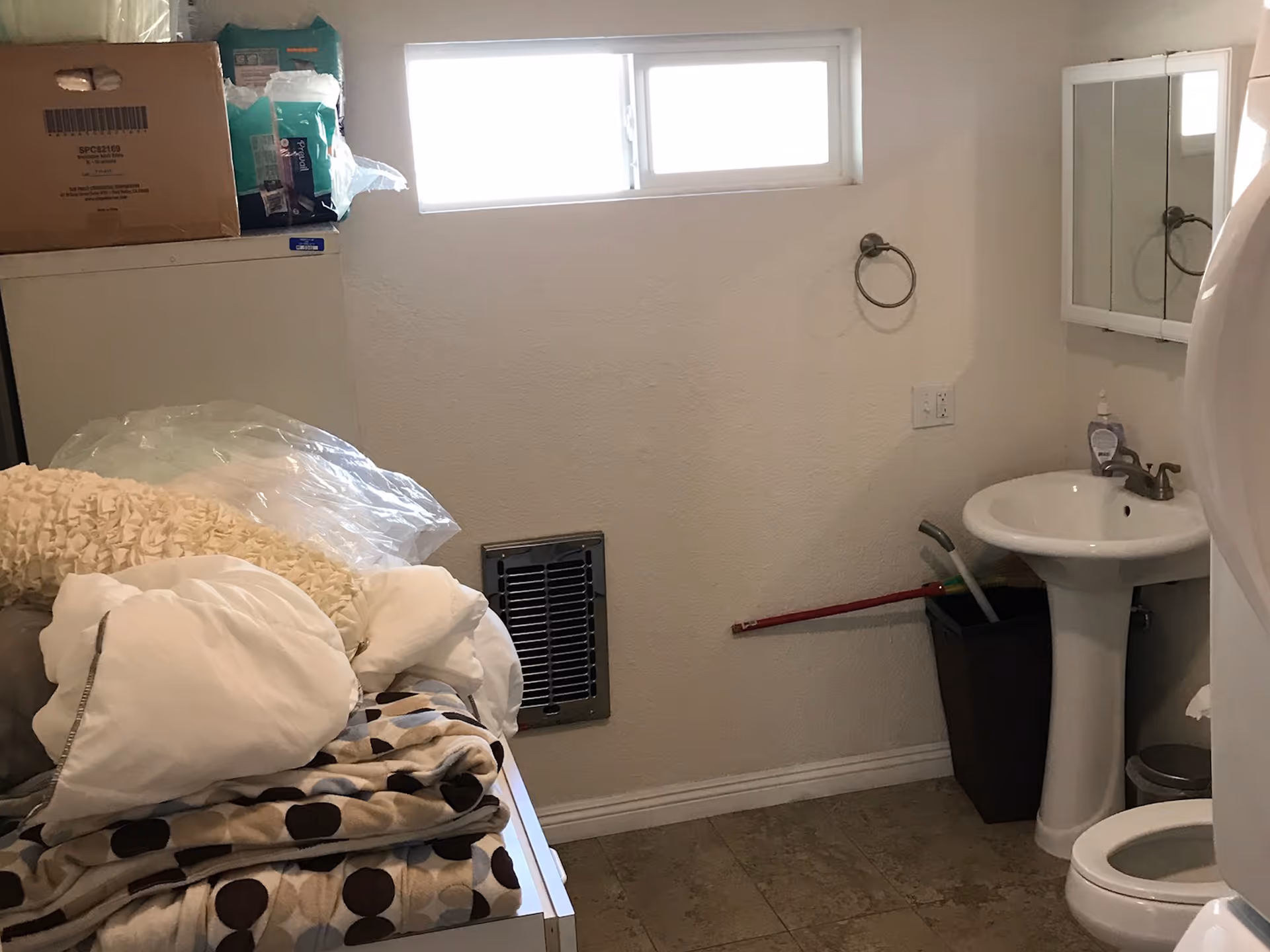 A small bathroom with a pedestal sink, a mirror cabinet above the sink, a toilet, and a window near the ceiling. There are various items including pillows, blankets, and boxes stacked on a surface to the left. A mop and trash bin are placed near the sink.