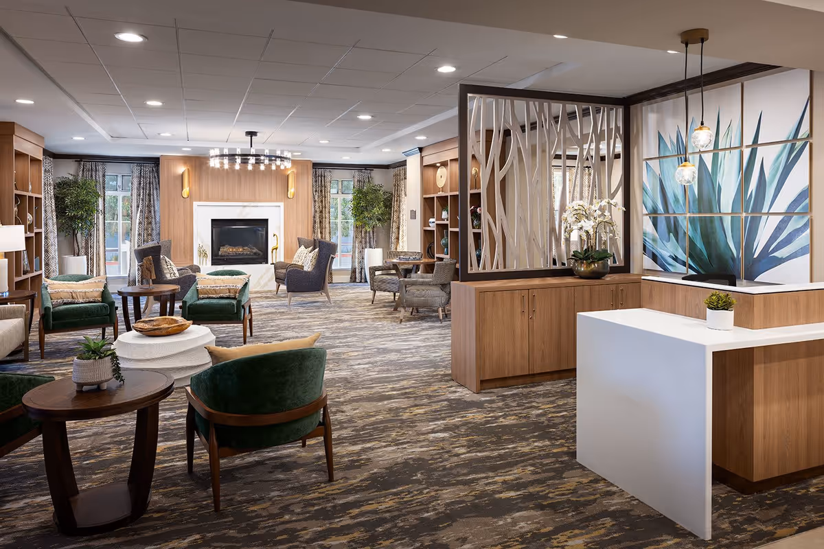 A spacious and well-lit senior living facility common area featuring comfortable seating with green and gray chairs, wooden tables, a modern fireplace with a wooden mantel, large windows with patterned curtains, decorative plants, and a reception desk with a large botanical artwork behind it.