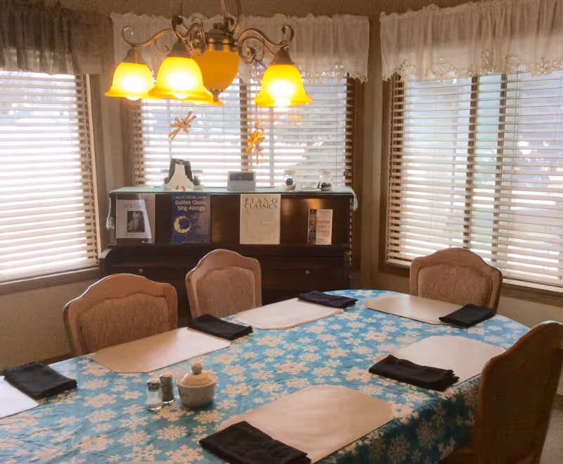 A dining room with a rectangular table covered with a blue tablecloth featuring white snowflake patterns. The table is set with beige placemats and black folded napkins at each seat. Four upholstered chairs surround the table. Behind the table is a piano with sheet music and books displayed on top. The room has large windows with wooden blinds and white lace curtains. A chandelier with five yellow lampshades hangs above the table.