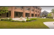 Exterior view of a brick senior living facility with multiple windows and a covered entrance. The foreground features a well-maintained lawn, landscaped bushes, and a paved walkway leading to the entrance.