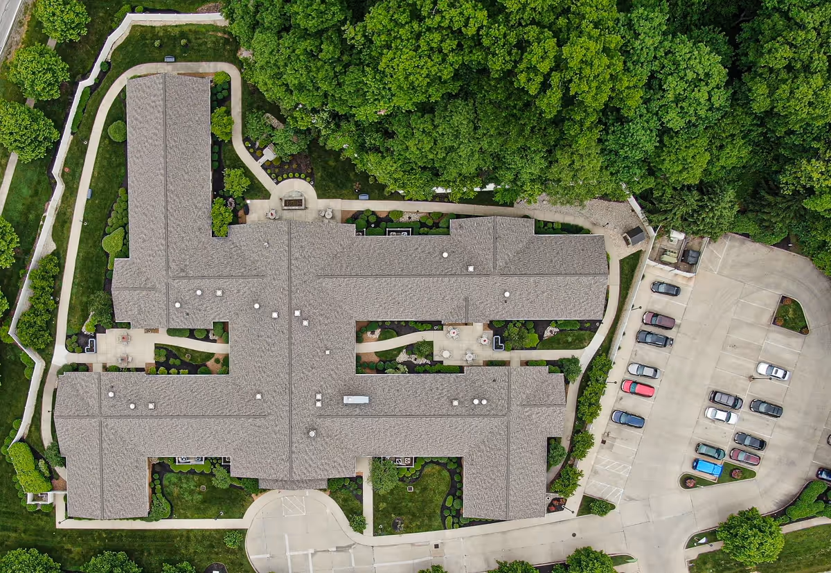 Aerial view of Arden Courts - ProMedica Memory Care Community building surrounded by green trees and landscaped gardens, with a parking lot on the right side.