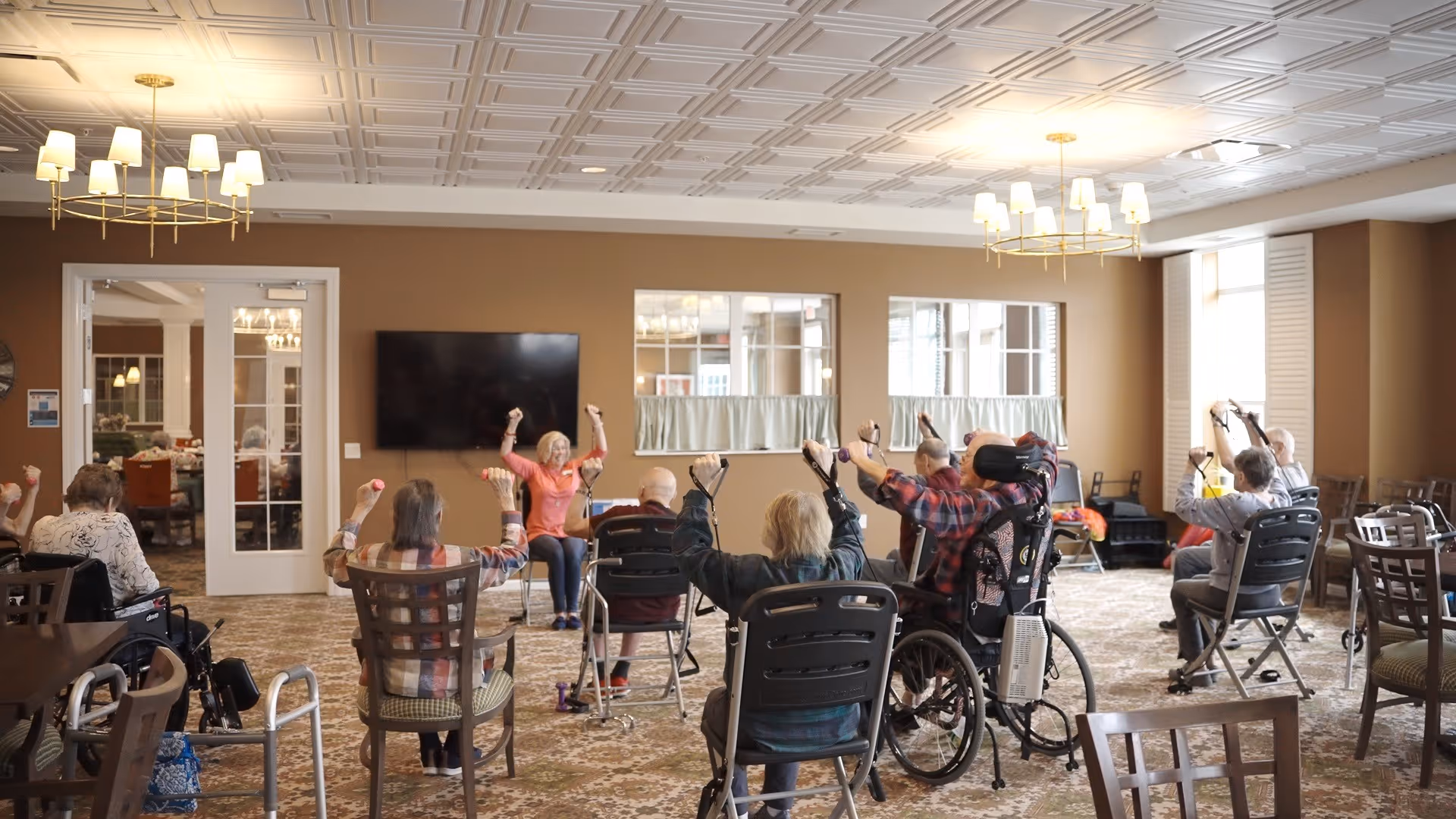 A group of elderly individuals seated in chairs and wheelchairs in a spacious room with beige walls and patterned carpet, participating in a seated exercise session led by an instructor at the front. The room has large windows, chandeliers, and a wall-mounted TV.