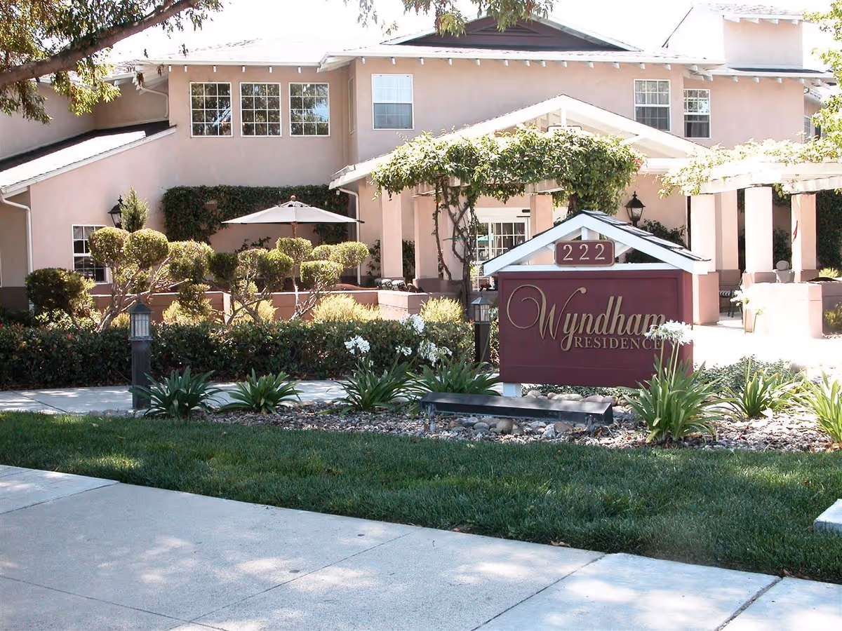 Exterior view of Wyndham Residence building with a landscaped garden in front, featuring trimmed bushes, flowers, and a sidewalk. A sign with the address number 222 and the name Wyndham Residence is prominently displayed near the entrance.