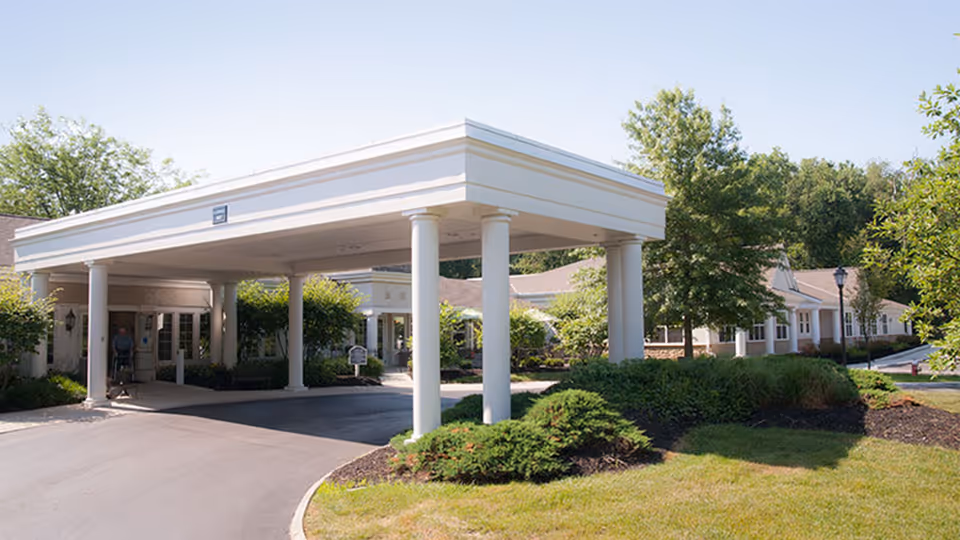 Entrance of New Albany - An Optalis Health & Rehabilitation Center featuring a covered drop-off area with white columns, surrounded by green shrubs and trees under a clear sky.