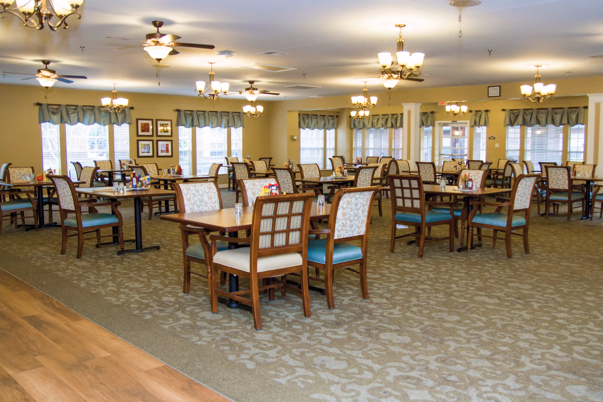 A spacious dining room with multiple wooden tables and chairs arranged neatly. The chairs have patterned upholstery and teal cushions. The room is well-lit with ceiling fans and chandeliers. Large windows with valances allow natural light to enter. Condiments and glasses are placed on the tables.