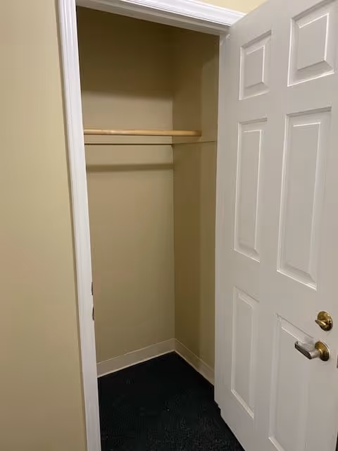 An empty closet with beige walls, a wooden hanging rod, and a white door partially open showing a brass-colored handle and lock.