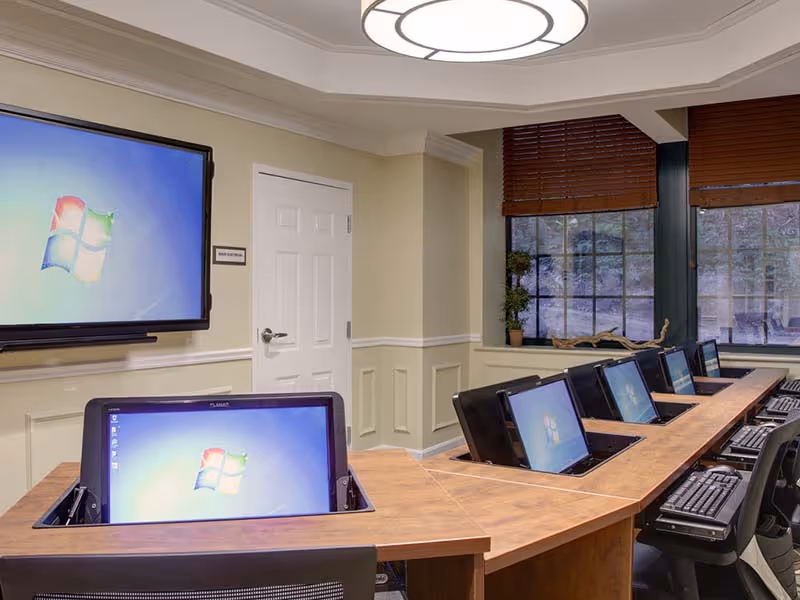 A computer lab with multiple desktop computers embedded in a wooden table, each displaying the Windows logo. There is a large wall-mounted screen also showing the Windows logo. The room has beige walls, a white door, large windows with brown blinds, and a modern circular ceiling light.