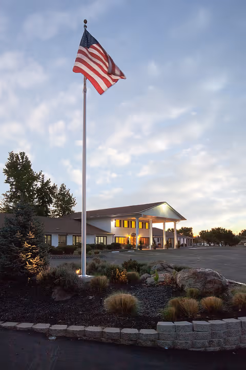 Exterior view of Westview Healthcare Center at dusk with an American flag on a tall flagpole in the foreground, landscaped garden with rocks and shrubs, and the building illuminated with warm lights.