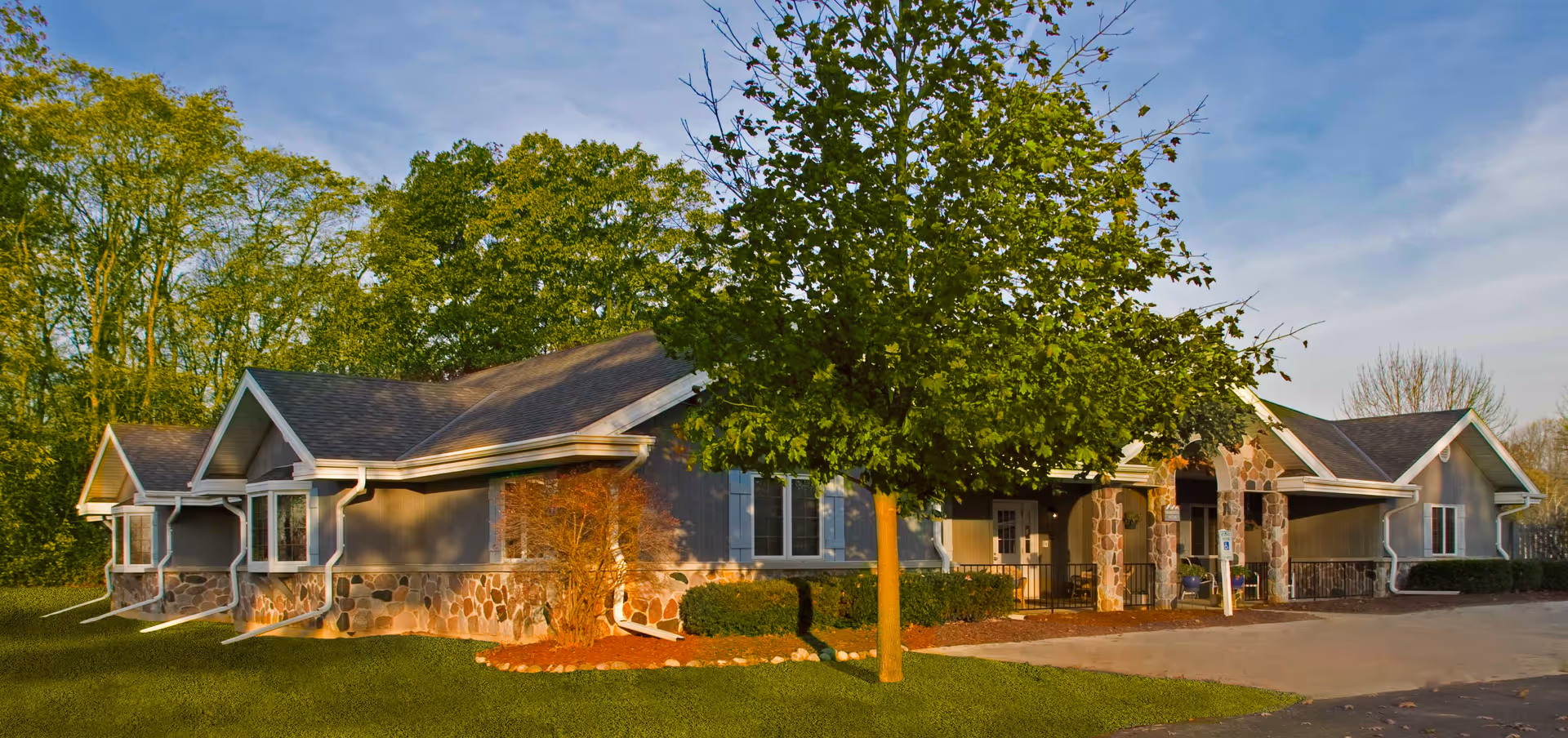 Exterior view of a single-story senior living facility building with stone and wood siding, surrounded by green trees and a well-maintained lawn under a partly cloudy sky.