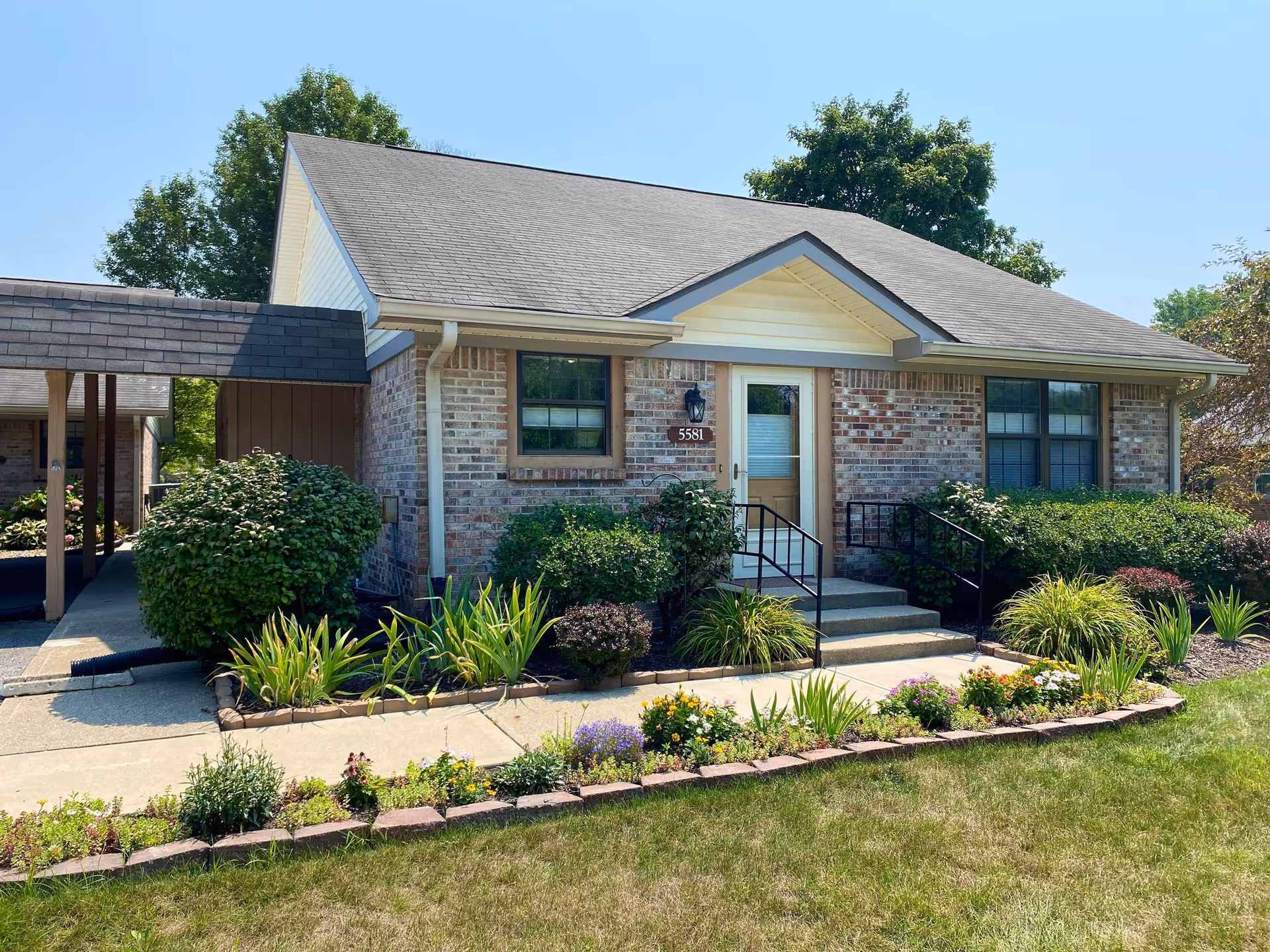 Exterior view of a single-story brick building with a gray shingled roof, a white door with a window, and two windows on either side. The building is surrounded by well-maintained landscaping including bushes, flowering plants, and a small garden bed bordered by bricks. A concrete walkway leads to the entrance with black handrails on either side of the steps. Trees are visible in the background under a clear blue sky.