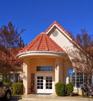 Front exterior view of a building with a red tiled roof and beige walls under a clear blue sky. The entrance features double glass doors with the number 715 above them, flanked by bushes and trees on either side. A handicap parking sign is visible near the entrance.
