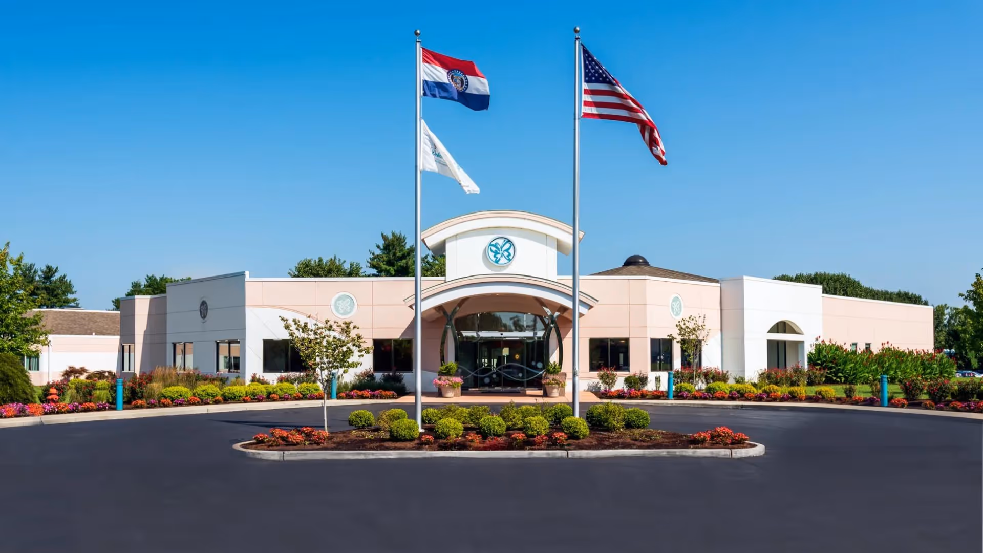 Front exterior view of Delmar Gardens South facility with a circular driveway, landscaped bushes and flowers, and three flagpoles displaying the Missouri state flag, an unidentified white flag, and the United States flag under a clear blue sky.