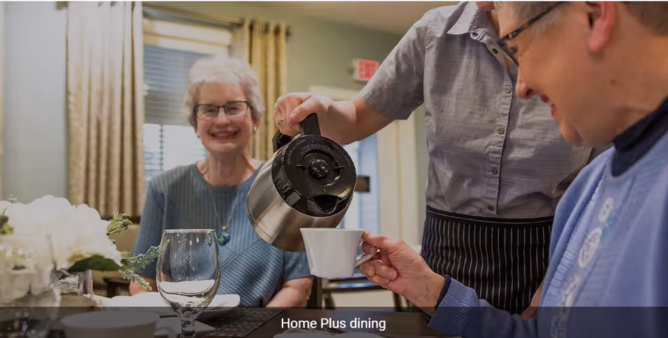 A smiling elderly woman sitting at a dining table while a staff member pours coffee into a cup held by another elderly woman in a cozy dining room setting.
