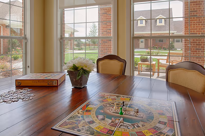 A wooden table with a board game set up on it and a box labeled 'Easy Handling' on the side. There is a small flower pot with white flowers in the center of the table. The table is surrounded by chairs with beige cushions. Large windows behind the table show a view of a brick building and outdoor seating area with benches and a small table.