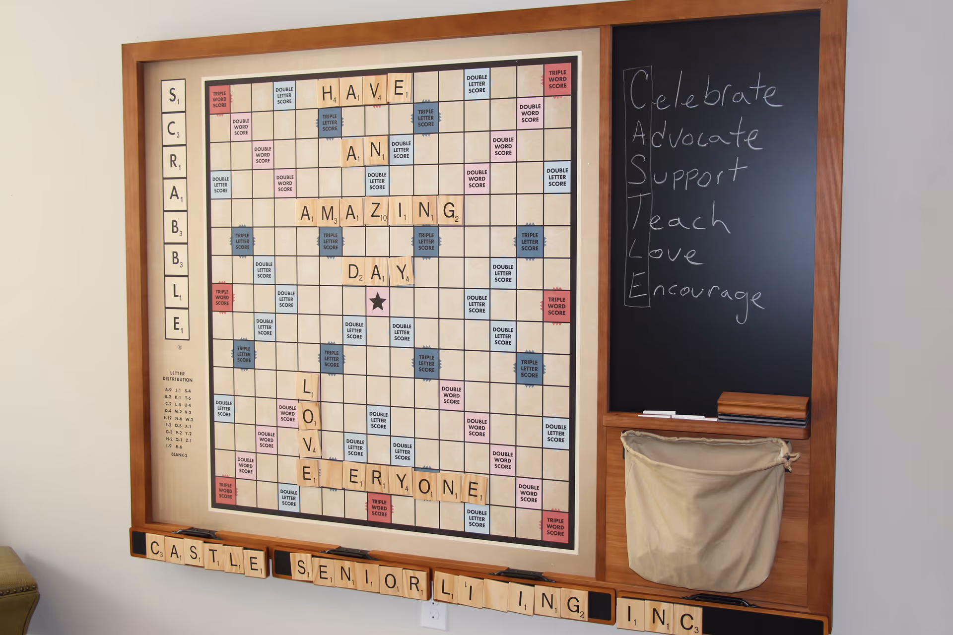 A large wooden-framed Scrabble board mounted on a wall with Scrabble tiles forming the phrase 'HAVE AN AMAZING DAY LOVE EVERYONE'. To the right of the board is a black chalkboard with the word 'CASTLE' written vertically and words Celebrate, Advocate, Support, Teach, Love, Encourage written horizontally next to each letter. Below the board are additional Scrabble tiles spelling 'CASTLE SENIOR LIVING INC.'. A beige cloth pouch hangs below the chalkboard.