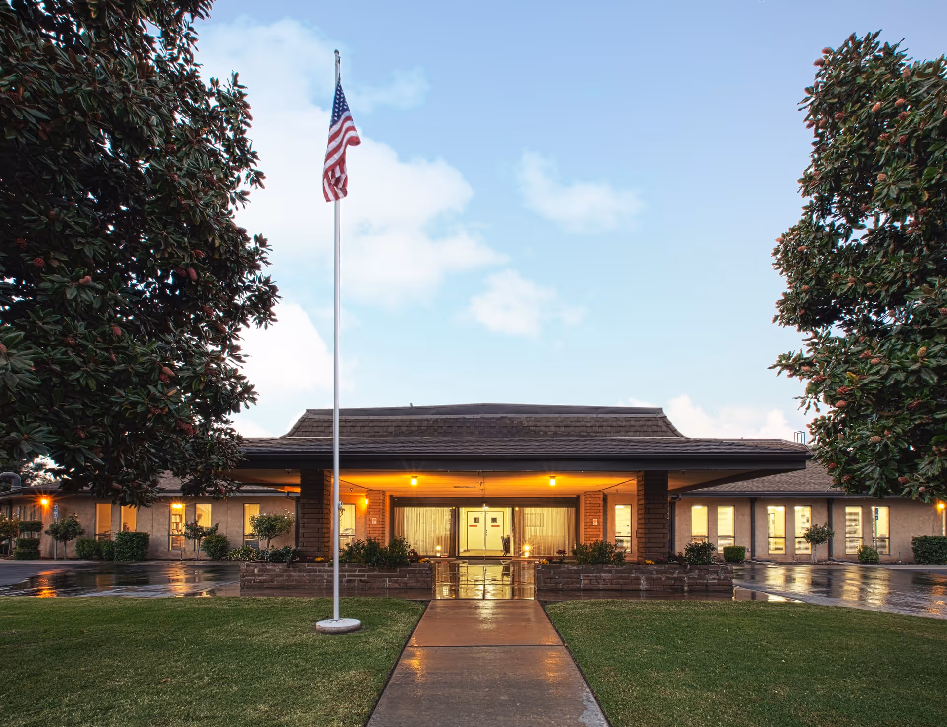 Front exterior view of Linwood Meadows Care Center building at dusk with an American flag on a flagpole in the foreground, flanked by two large trees and a wet walkway leading to the entrance.