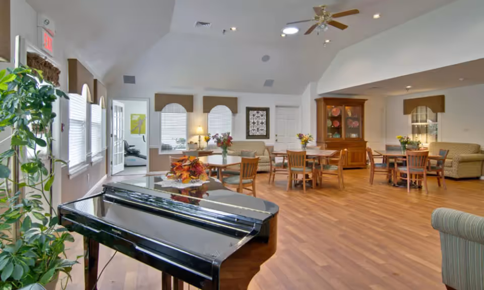 A spacious and well-lit common area with wooden flooring, several tables and chairs, a black grand piano in the foreground, a large plant near the windows, and comfortable seating including sofas. The room has high ceilings with ceiling fans and recessed lighting, and there are decorative elements such as flowers on the tables and framed artwork on the walls.