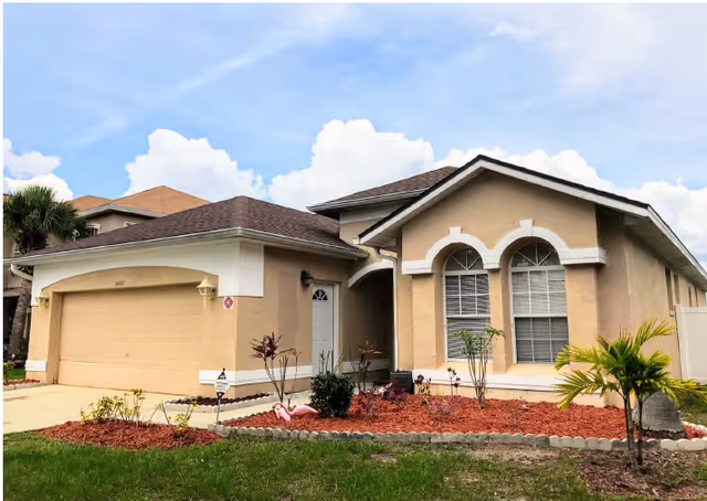 Single-story beige house with a brown roof, a two-car garage, and a small landscaped front yard with plants and a pink flamingo decoration under a partly cloudy sky.