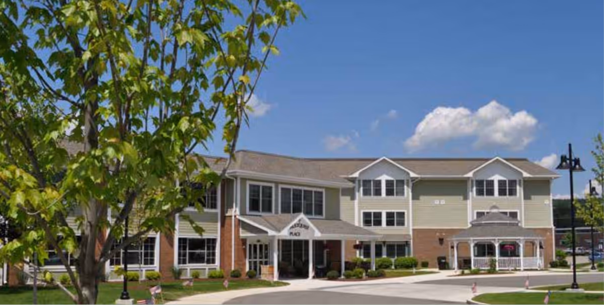 Exterior view of a two-story senior living facility named Hultquist Place with a covered entrance, a white gazebo, and a tree with green leaves in the foreground under a blue sky with some clouds.