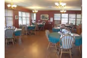 Dining room with multiple round tables covered with green tablecloths, surrounded by wooden chairs with cushions. The room has large windows with blinds, wooden flooring, and chandeliers hanging from the ceiling. A painting and a sideboard are visible against the far wall.