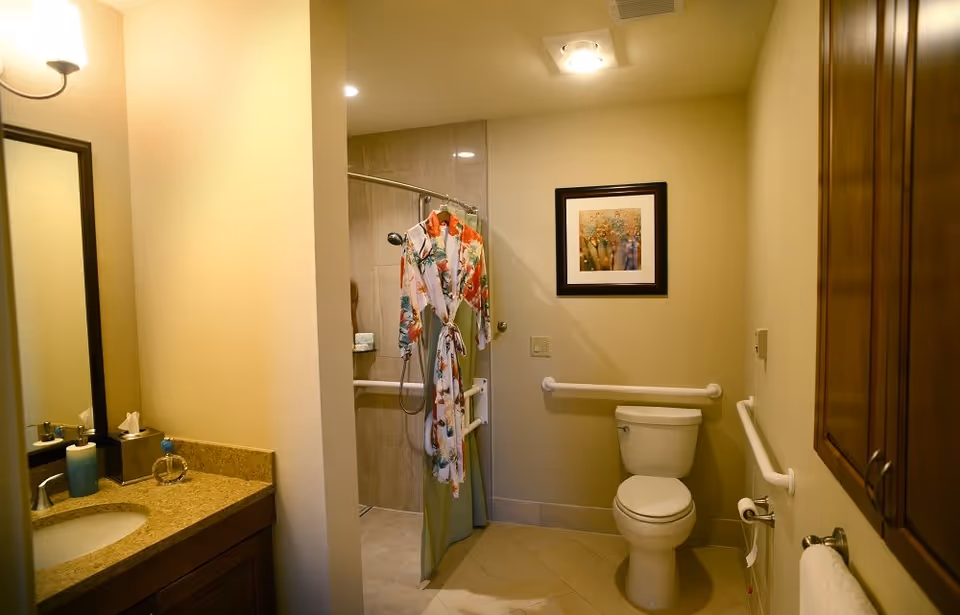 A bathroom with beige walls and tiled floor featuring a toilet with grab bars on the walls, a walk-in shower with a floral robe hanging on the shower curtain rod, a sink with a granite countertop, a mirror, and a framed floral picture on the wall.