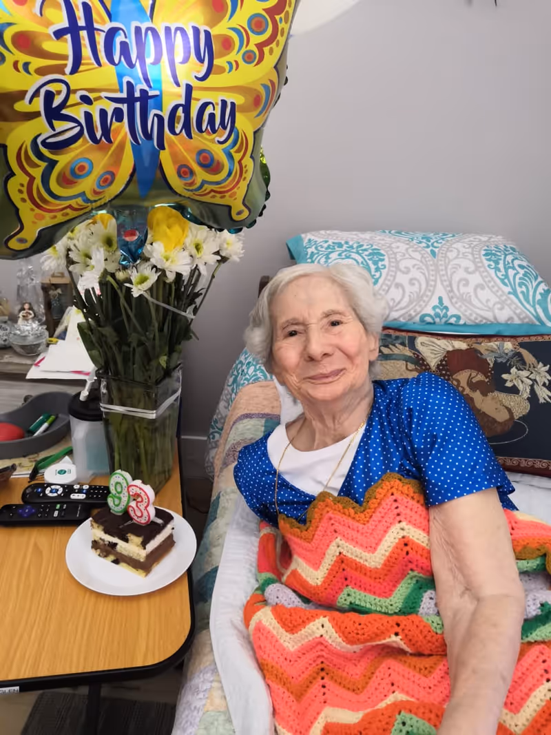 An elderly woman with white hair lying in bed covered with a colorful crocheted blanket, smiling. Next to her is a table with a slice of birthday cake with candles showing the number 93, a vase of white and yellow flowers, and a large butterfly-shaped balloon that says 'Happy Birthday'.