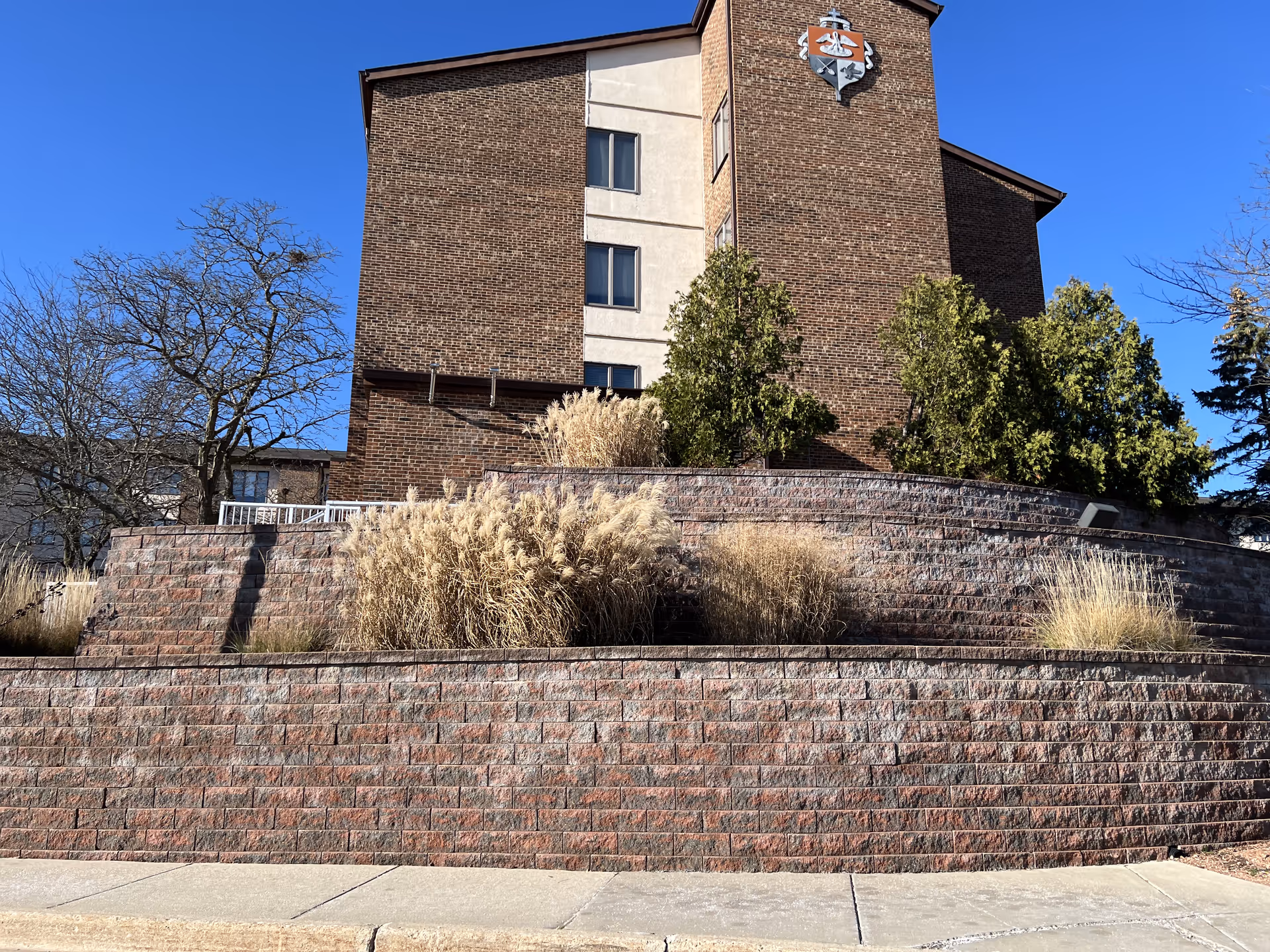 Exterior view of a multi-story brick building with a crest emblem near the top. In front of the building is a tiered retaining wall made of red bricks with ornamental grasses and shrubs planted on each level. The sky is clear and blue.
