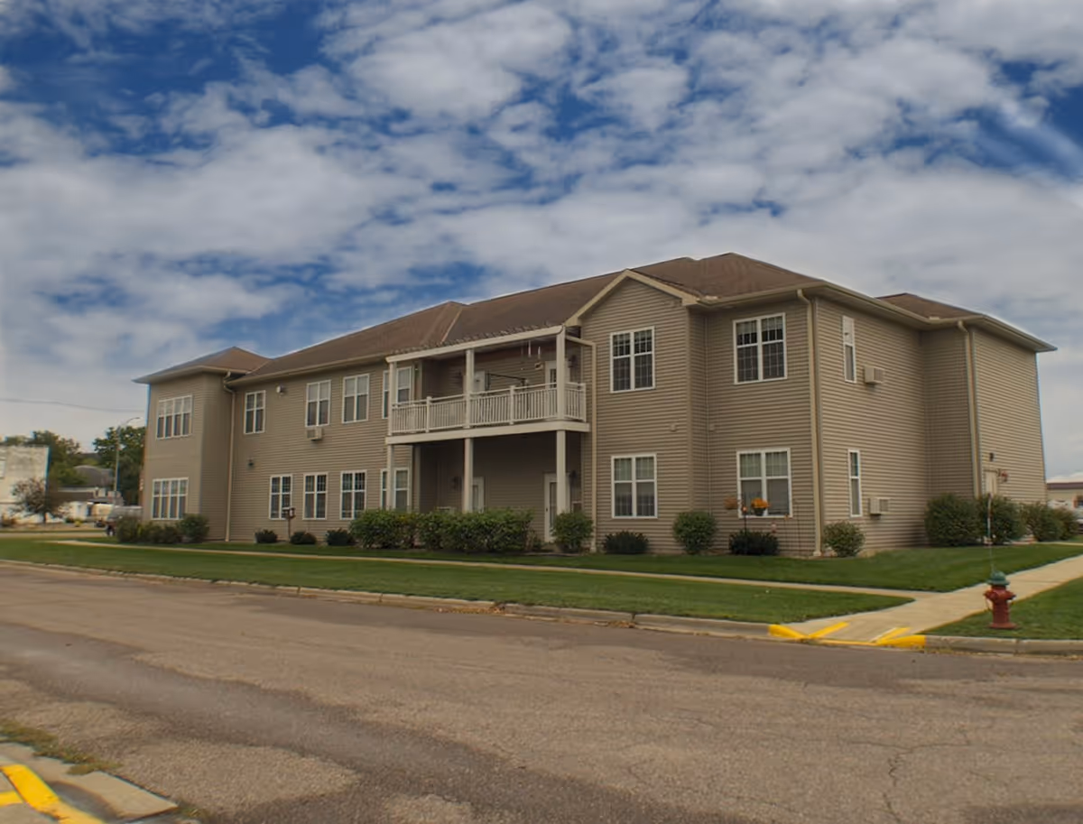 Exterior view of a two-story senior living facility building with beige siding, multiple windows, a small balcony, and a well-maintained lawn under a partly cloudy sky.