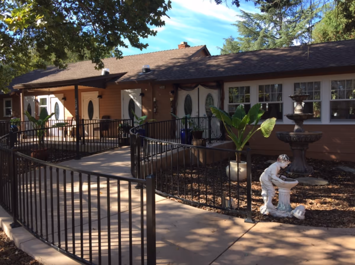 Exterior view of a single-story building with a brown roof and beige walls, featuring multiple white doors with oval glass panels. A black metal railing lines a curved concrete walkway leading to the entrances. There are large green potted plants and a decorative statue of a child drinking from a birdbath near a multi-tiered fountain. Trees with green leaves provide shade over the area.