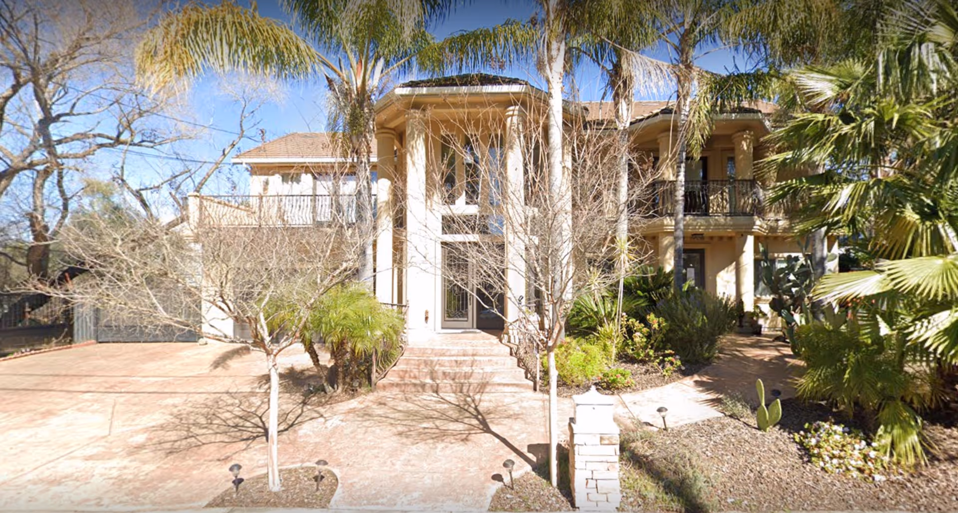 Front exterior view of a large, two-story house with tall columns, a balcony, and a landscaped yard featuring palm trees and other plants under a clear blue sky.