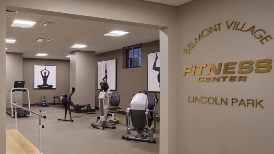 Interior view of the Belmont Village Senior Living Lincoln Park fitness center featuring exercise equipment including a treadmill, rowing machine, stationary bikes, and free weights. The room has neutral-colored walls with framed artwork of yoga poses and a window letting in natural light.