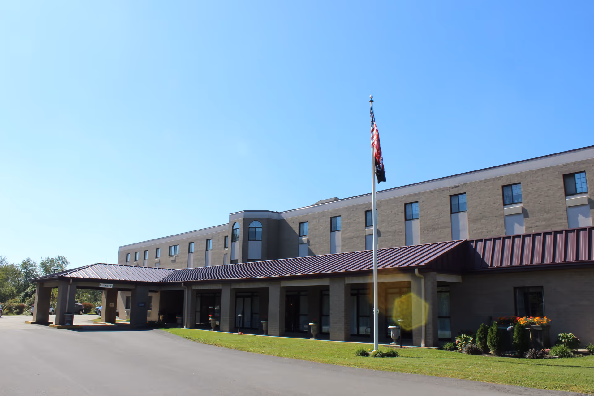 Exterior view of a multi-story senior living facility building with a covered entrance and an American flag on a flagpole in front. The building has multiple windows and a brown roof, with a clear blue sky overhead.