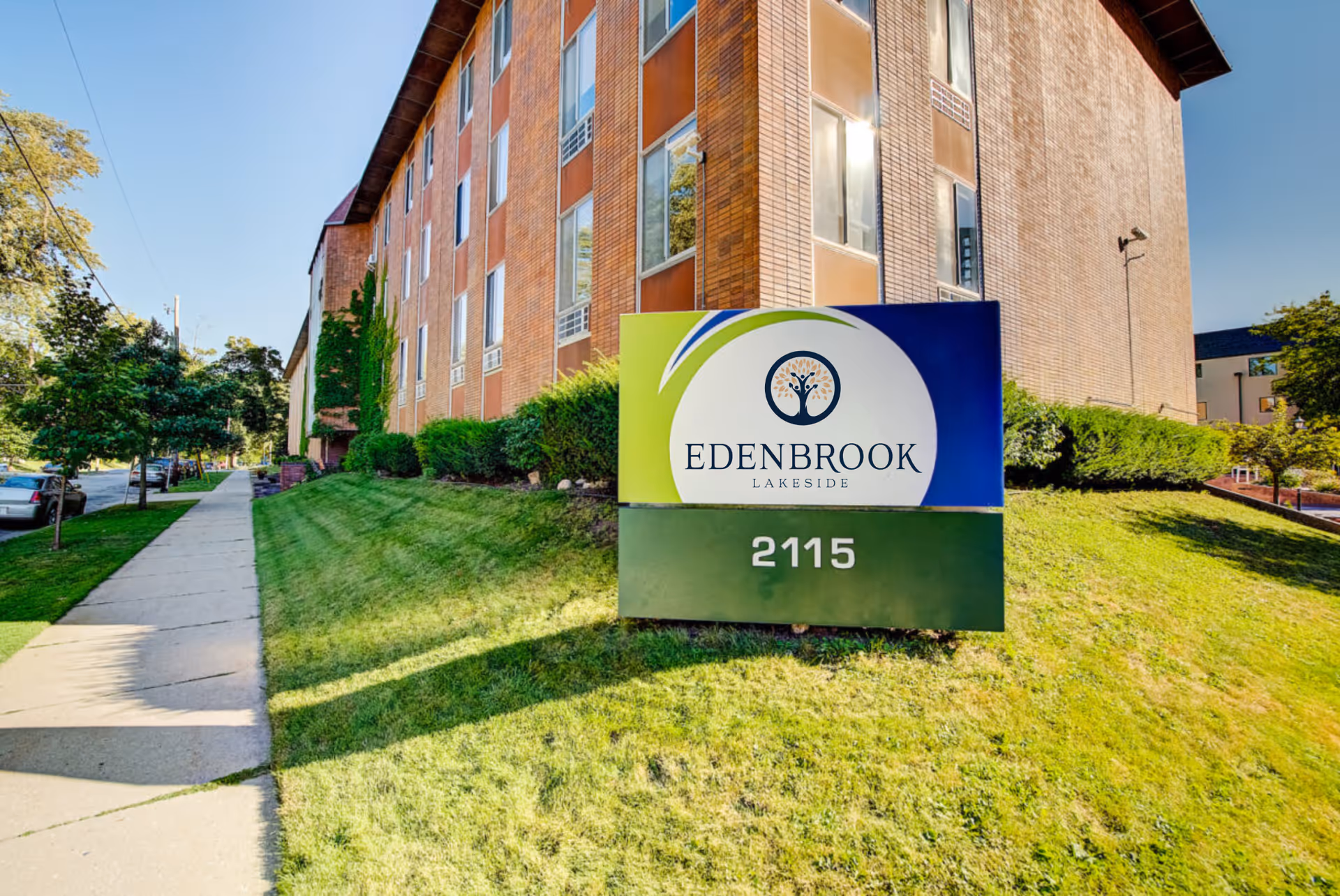 Exterior view of a multi-story brick building with a green lawn and sidewalk. In front of the building is a sign that reads 'Edenbrook Lakeside' with the number 2115 below it. Trees and parked cars are visible along the sidewalk.