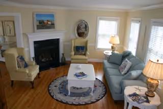 A cozy living room with a light blue sofa, two beige armchairs, a white coffee table on a round rug, a fireplace with a painting above it, and two table lamps providing warm lighting.