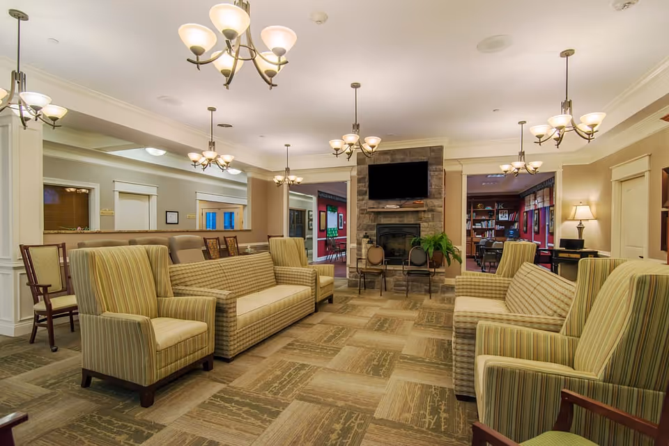 A spacious and well-lit living room area in a senior living facility featuring multiple striped armchairs and sofas arranged around a central stone fireplace with a mounted flat-screen TV above it. The room has beige walls, carpeted flooring with a patterned design, and several ceiling light fixtures providing warm lighting. There are doorways leading to other rooms, including a room with bookshelves and framed pictures on the walls.