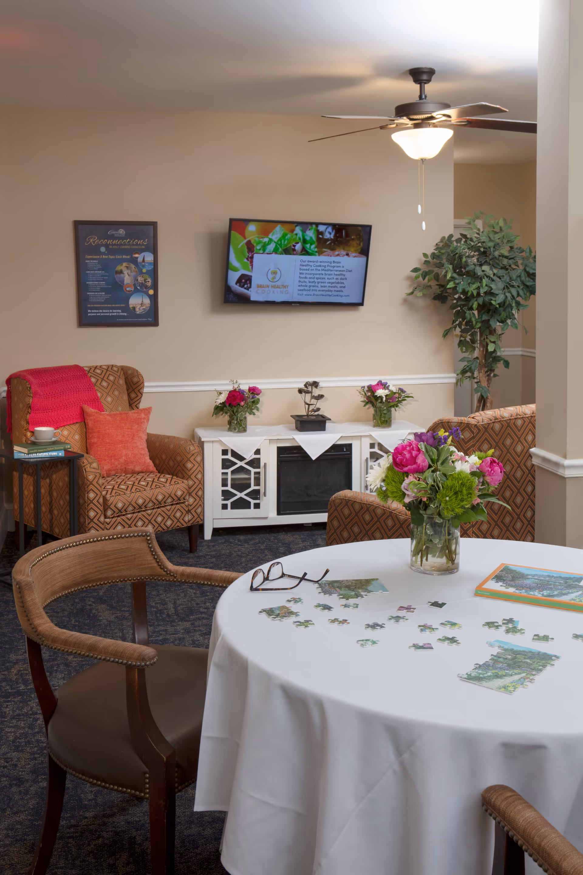 A cozy living room area with a round table covered with a white tablecloth, a partially completed jigsaw puzzle, and a vase of flowers. There are two wooden chairs around the table. In the background, there are two patterned armchairs with cushions, a small side table with a cup and books, a white cabinet with decorative items and flowers, a flat-screen TV mounted on the wall, a framed picture, and a ceiling fan with a light. A tall green plant is in the corner.