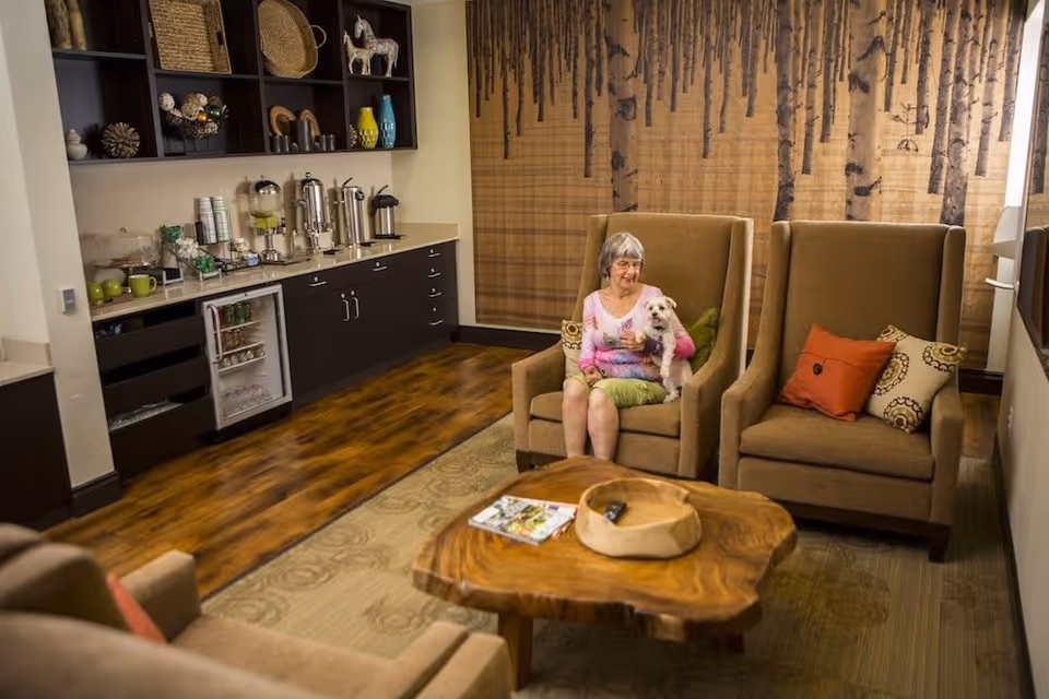 A cozy living room area with wooden flooring and a nature-themed wall mural. An elderly woman is sitting in a high-back armchair holding a small white dog. The room features a wooden coffee table with a magazine and a decorative bowl, two armchairs with colorful cushions, and a counter with a mini fridge, coffee urns, cups, and decorative items on shelves.