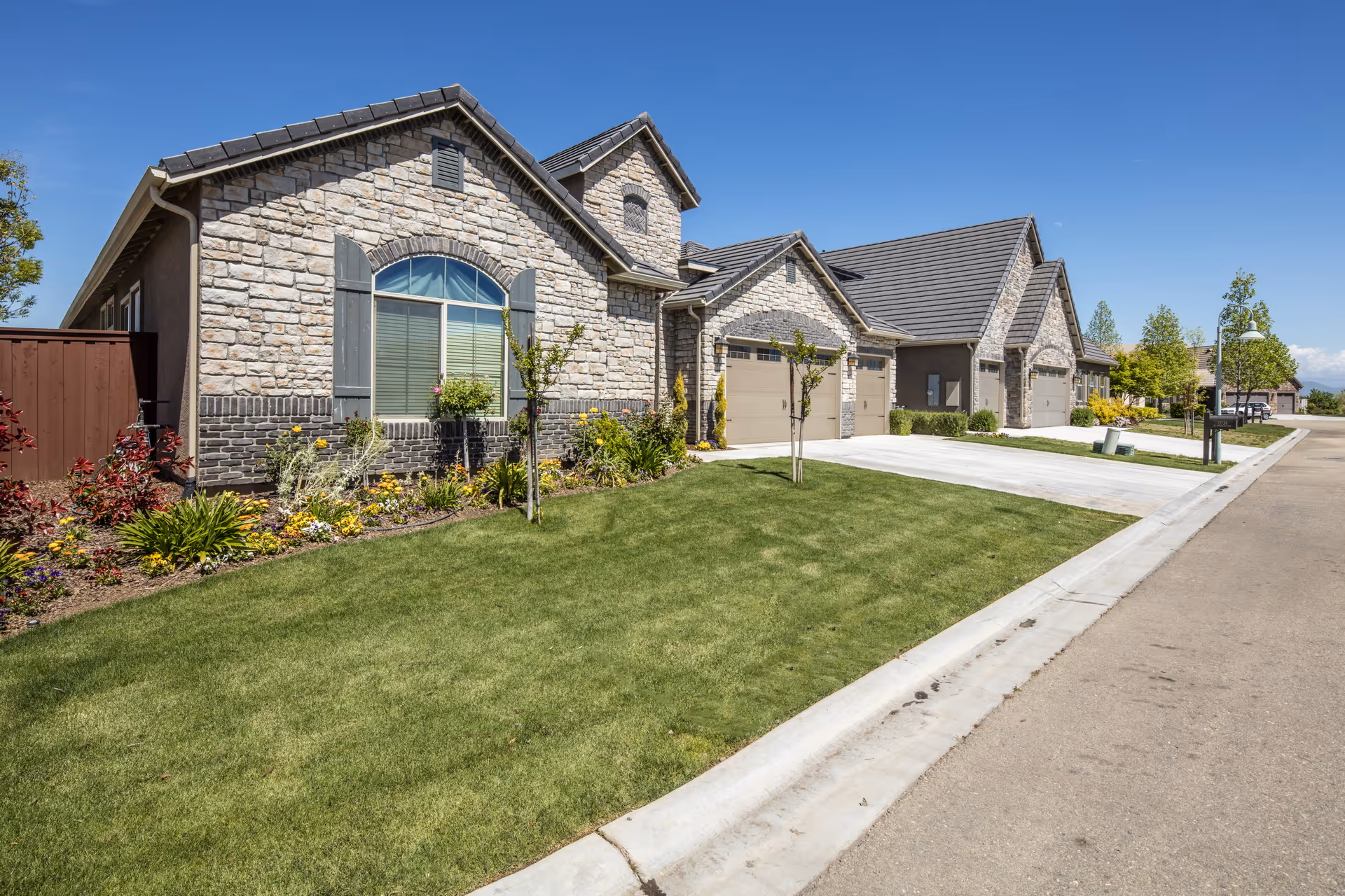 Exterior view of a residential care facility showing a row of stone and brick houses with garages, well-maintained green lawns, small trees, and flower beds under a clear blue sky.