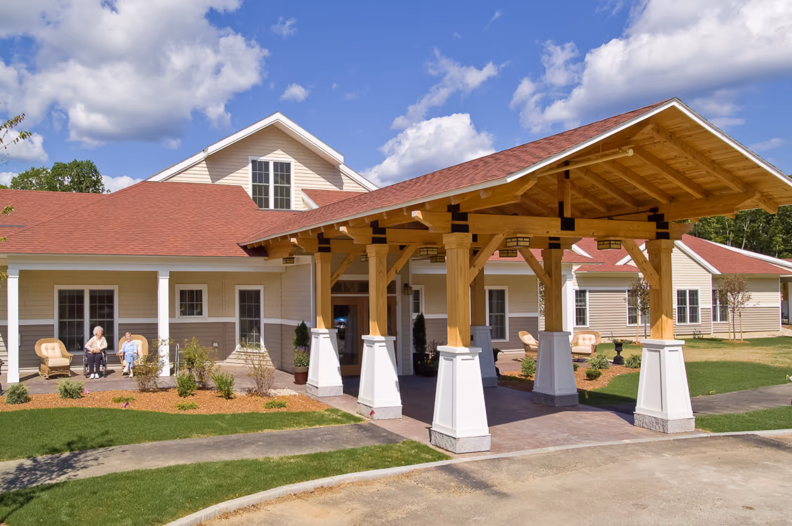 Exterior view of a senior living facility with a large wooden covered entrance. Two elderly women are sitting on chairs on the porch near the entrance. The building has beige siding with white trim and a red roof. The sky is blue with some clouds.