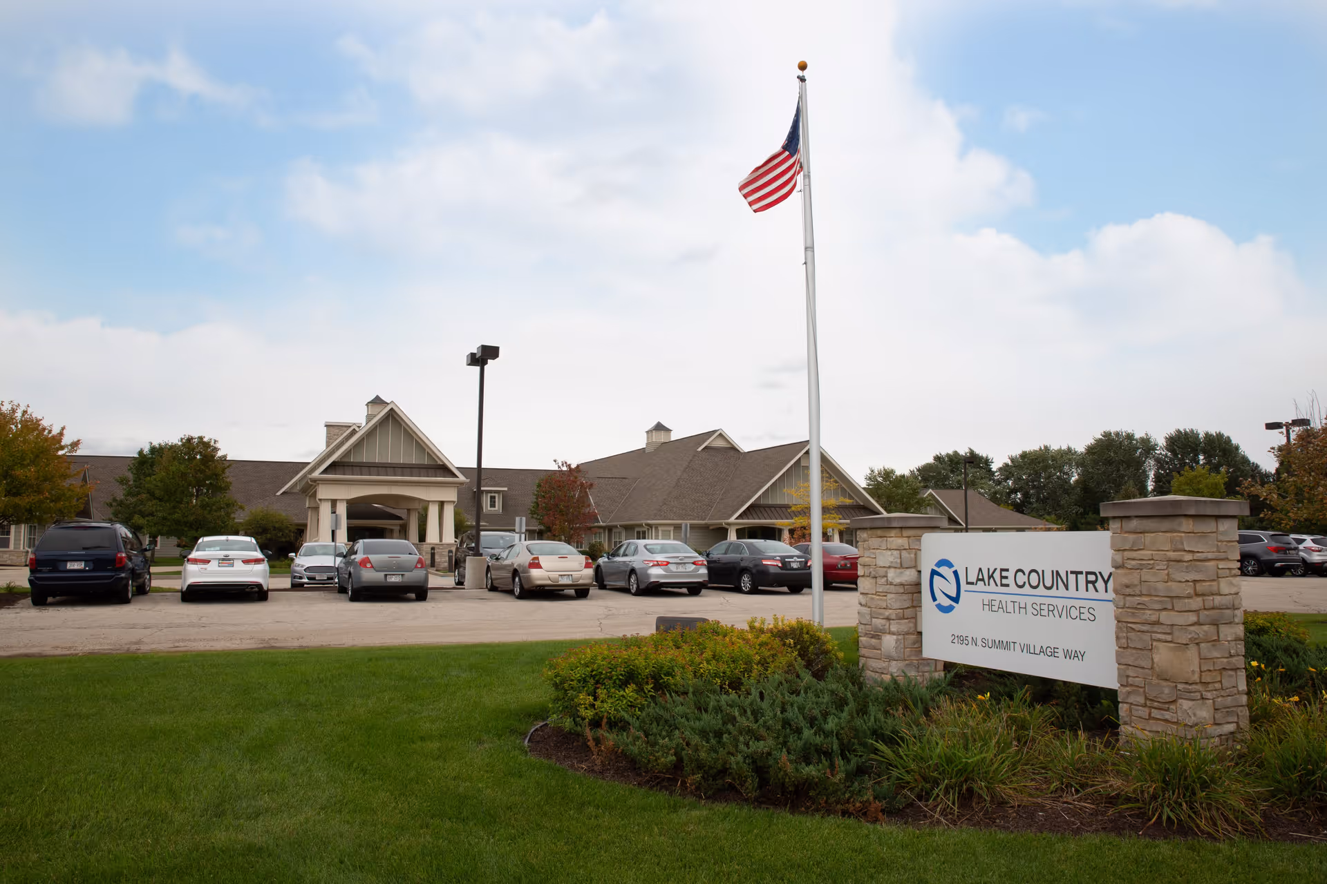Exterior view of Lake Country Health Services building with a parking lot in front, several parked cars, a flagpole with an American flag, and a sign displaying the facility's name and address surrounded by greenery.