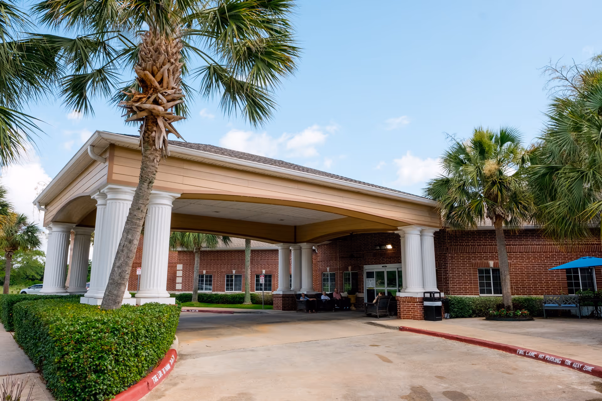 Entrance of a senior living facility with a covered drop-off area supported by white columns, surrounded by palm trees and neatly trimmed hedges. The building is made of red brick and has large windows. There are a few people sitting under the covered area near the entrance.