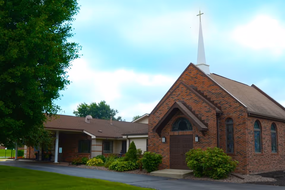 Exterior view of a brick building with a tall white steeple topped with a cross, surrounded by green bushes and trees under a partly cloudy sky.