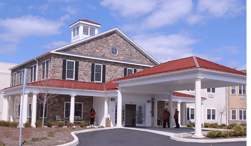 Exterior view of a senior living facility building with stone and siding walls, black shutters, and a red metal roof. The entrance features a covered drop-off area supported by white columns. Three people are standing near the entrance. The sky is partly cloudy.