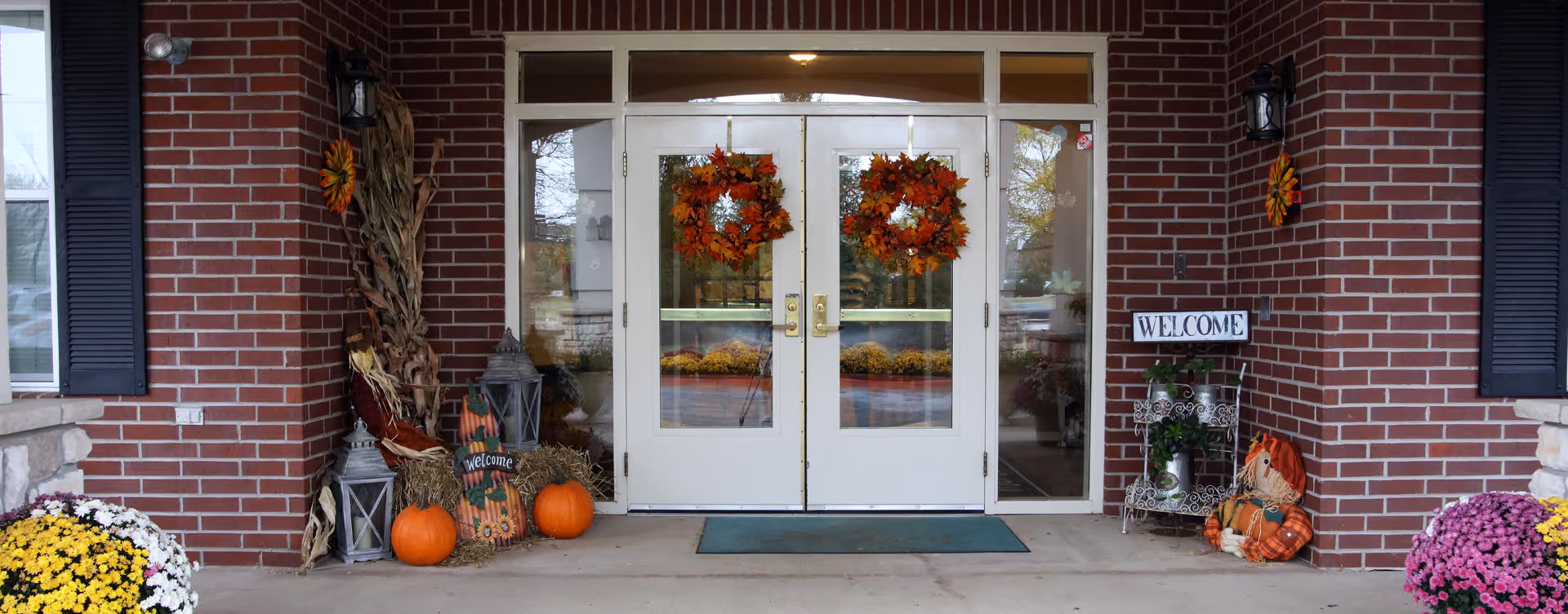 Entrance of a brick building decorated for autumn with two white double doors adorned with orange and red leaf wreaths. On either side of the doors are fall decorations including pumpkins, corn stalks, lanterns, a scarecrow, and potted flowers. A welcome sign is displayed on the right side near the door.