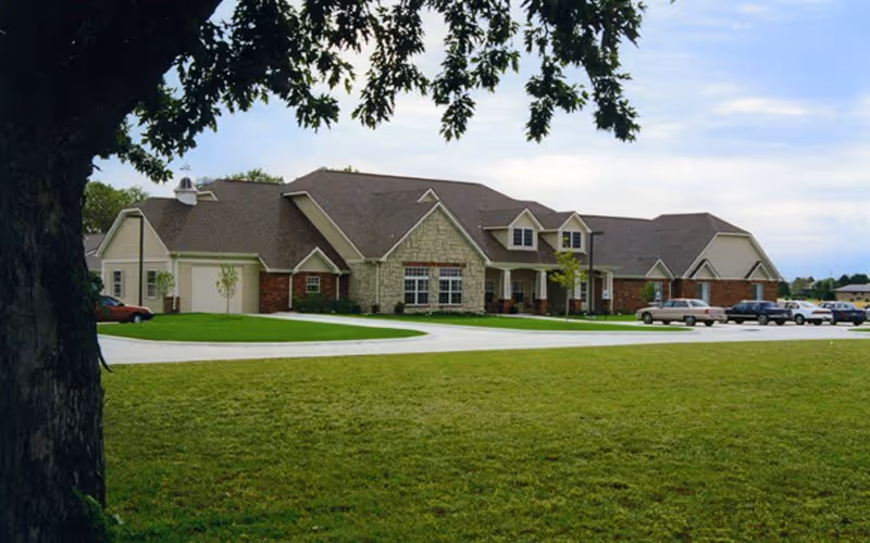 Exterior view of a single-story senior living facility building with a combination of brick and stone facade, multiple windows, and a sloped roof. The building is surrounded by a well-maintained lawn and a paved driveway with several parked cars. A large tree is visible in the foreground on the left side.