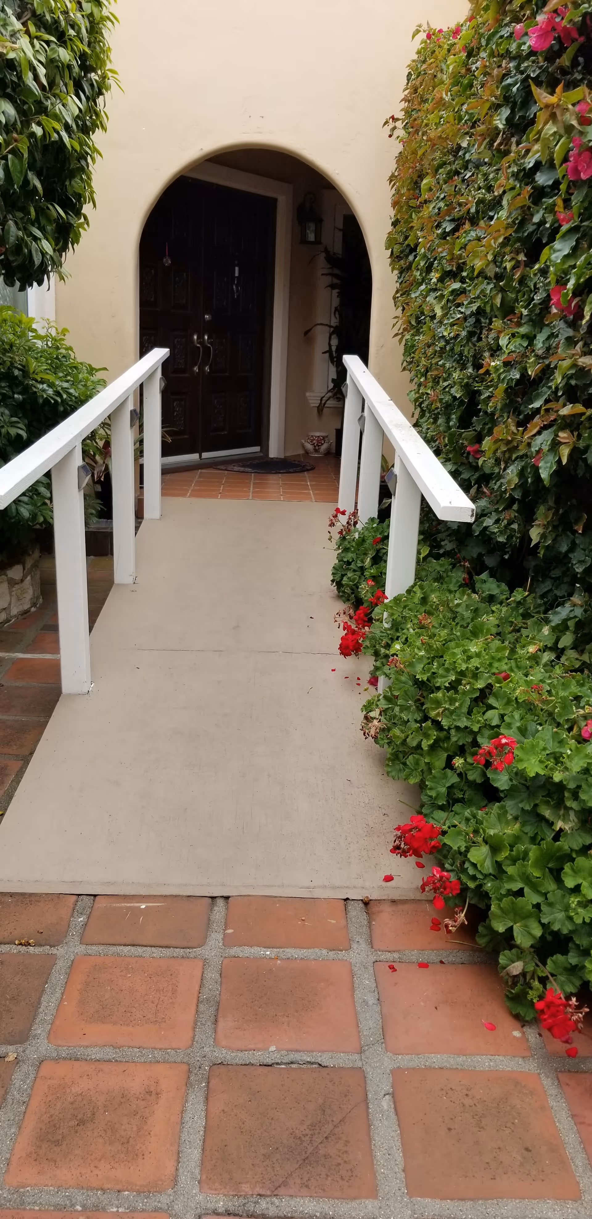 A concrete ramp with white handrails on both sides leading to a dark wooden double door entrance set in a beige stucco wall with an arched doorway. The ramp is flanked by green bushes and red flowers on the right side and a leafy tree on the left side. The ground in front of the ramp is tiled with reddish-brown square tiles.